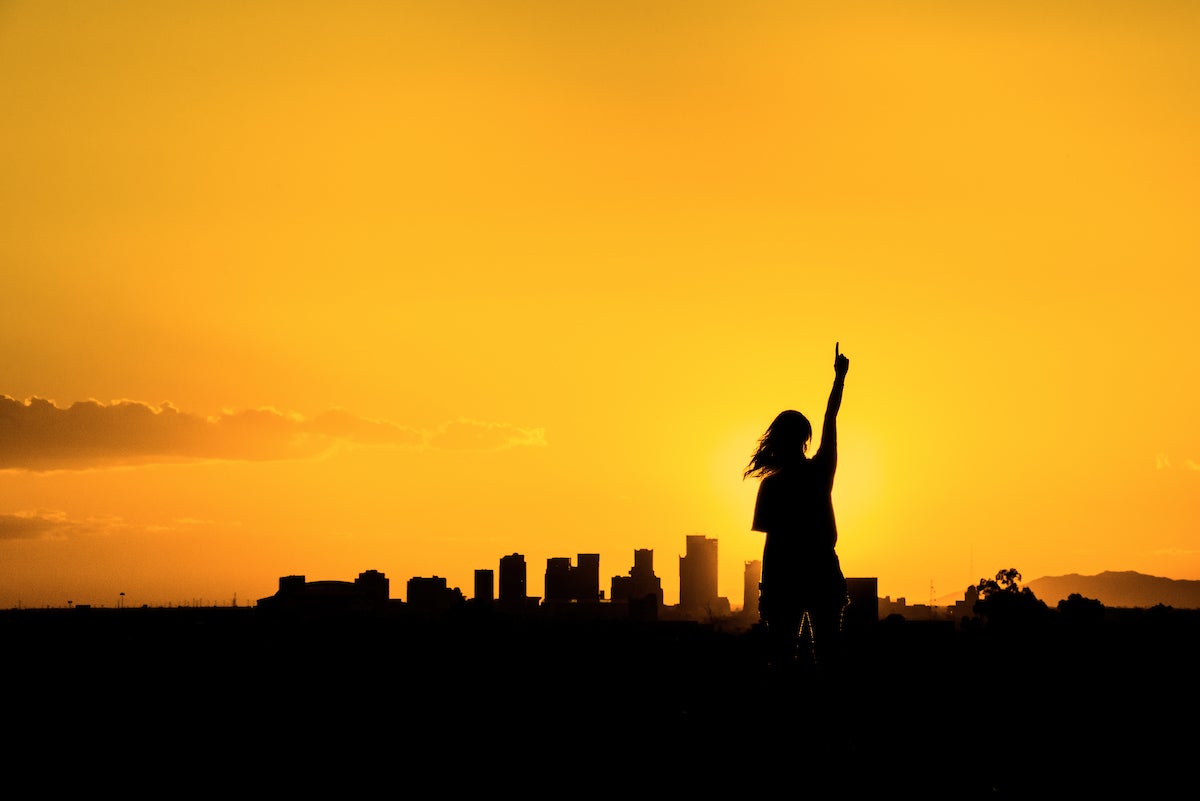 A person in shadow stands at sunset in front of a shadowed city skyline making the "number one" gesture with her left hand.