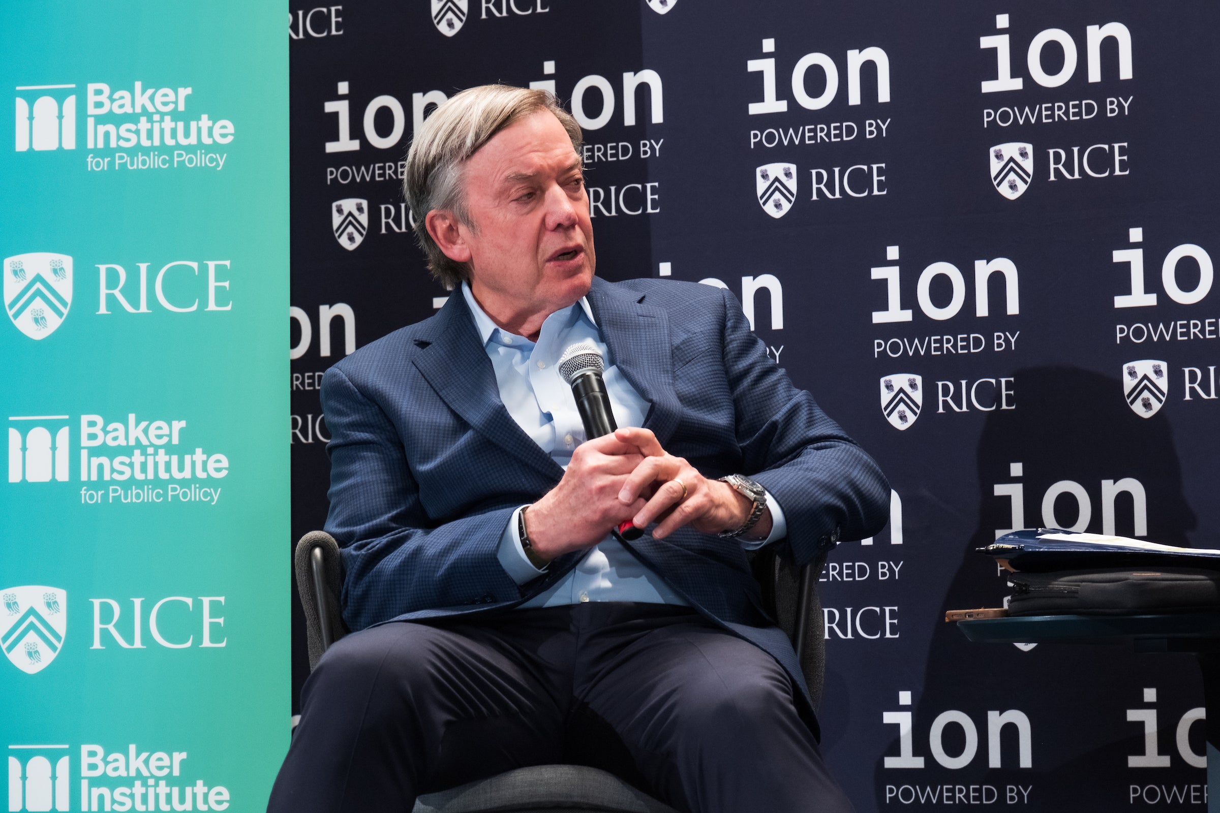 A person in business attire sits and speaks into a microphone on-stage during a fireside chat about US research universities.  