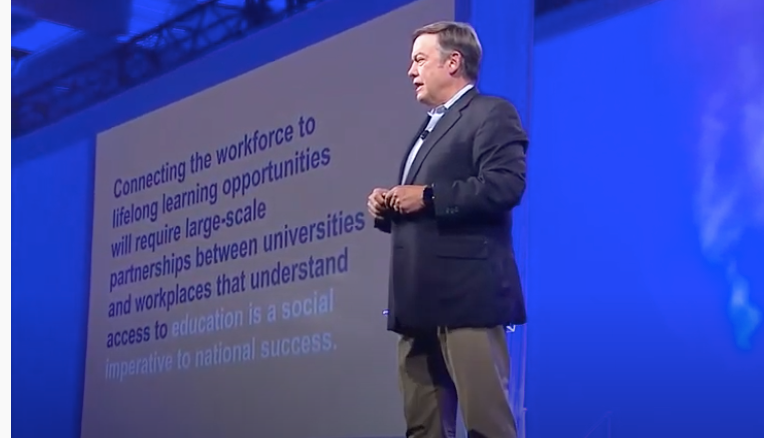 A middle-aged man in beige slacks and a blue jackets stands on a stage in front of a PowerPoint slide at the 2019 ASU GSV Summit.