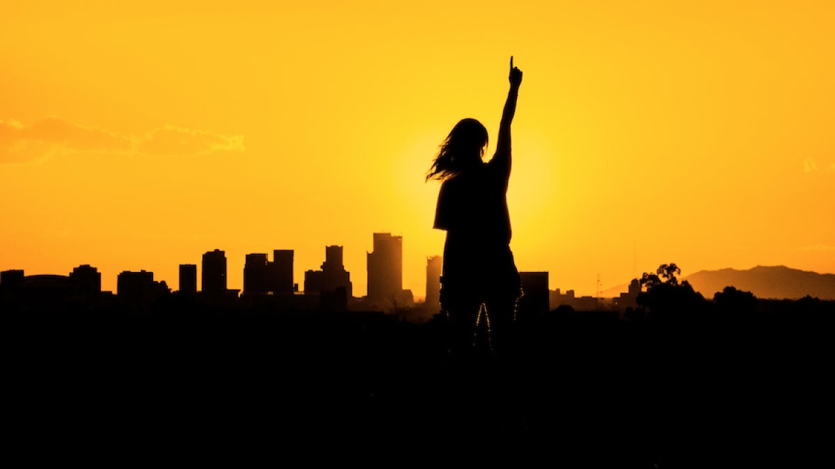 A person in shadow stands at sunset in front of a shadowed city skyline making the "number one" gesture with her left hand. 