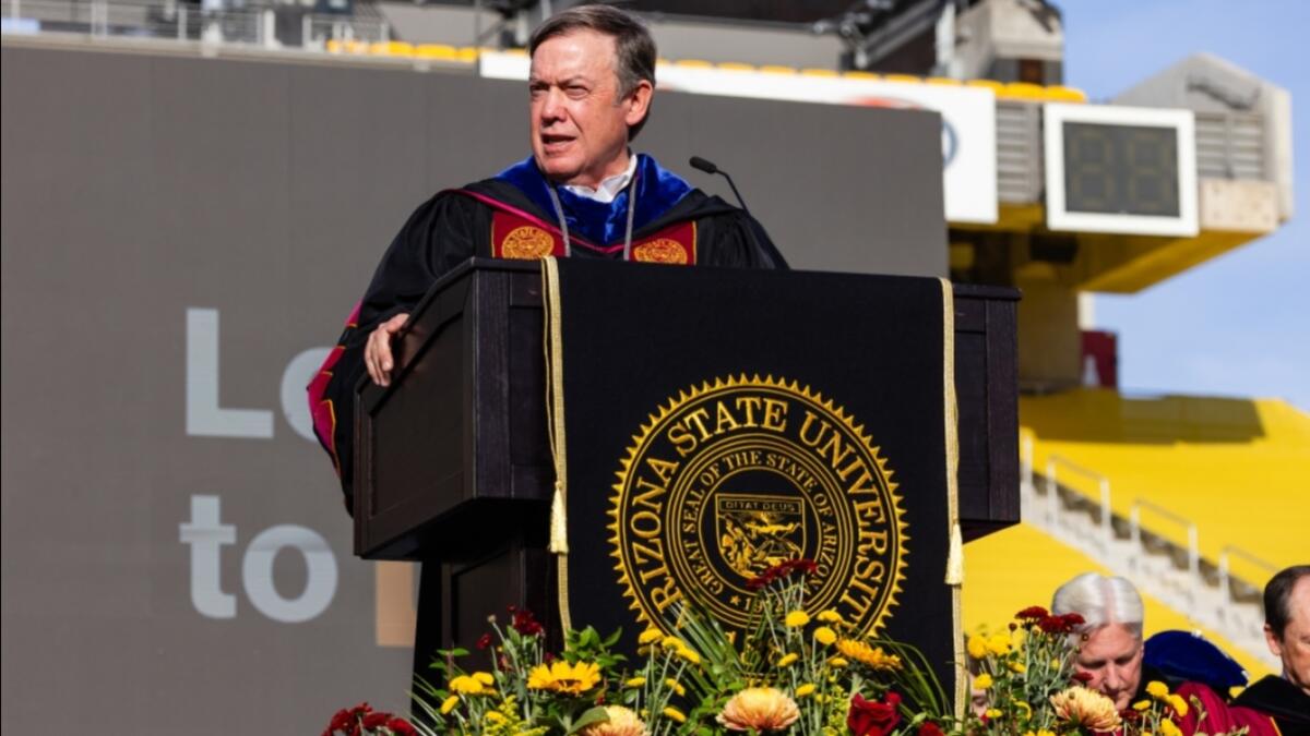 A university president stands at a podium on stage at an outdoor stadium while giving remarks at a university commencement ceremony. 