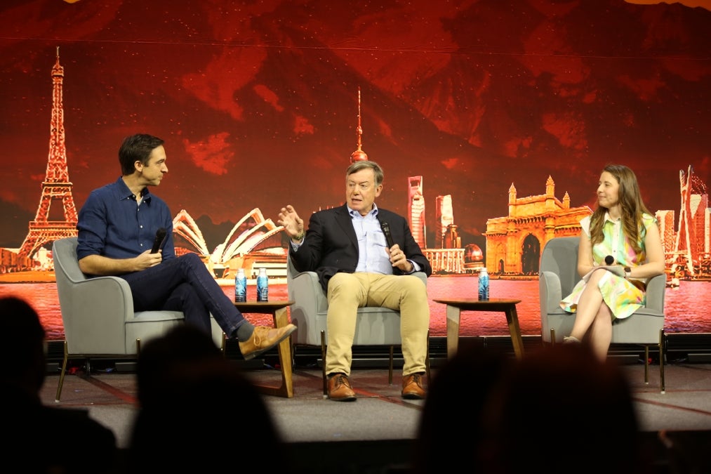 Three people sit on-stage un front of a backdrop featuring famous buildings from around the world while discussing learning through games. 