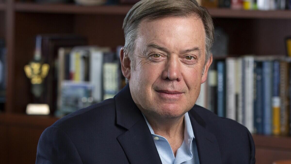 A head shot of a person in a blue shirt and jacket seated in front of book shelves in an office.  