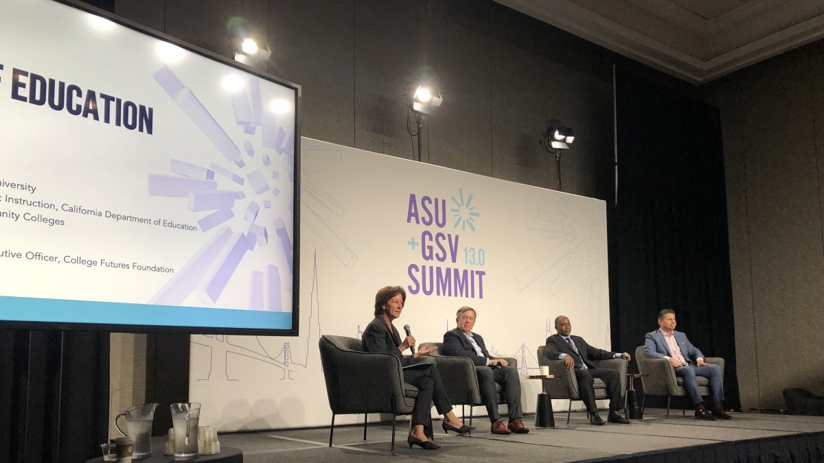 Four adults in business attire sit on a raised stage next to a large monitor and in front of a backdrop that reads "ASU + GSV Summit" in purple lettering. The first person on the panel is speaking into a microphone. 