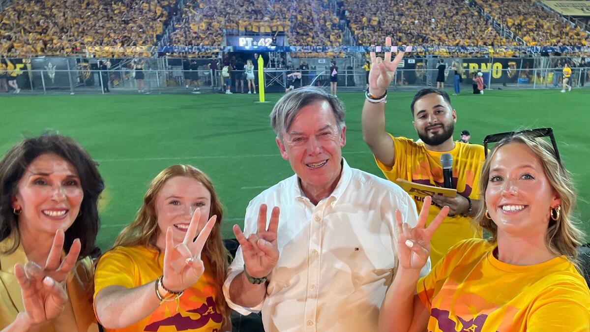 Two university administrators and three students stand smiling and making pitchfork hand gestures in front of thousands of first-year college students.  