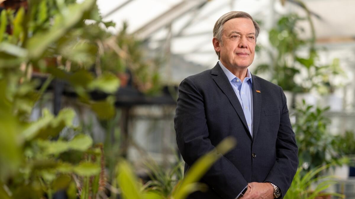 A person in a blue shirt and blazer stands smiling in a university greenhouse. 