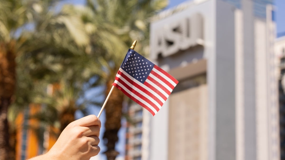 A hand holds a small American flag outside in front of a large ASU sign. 