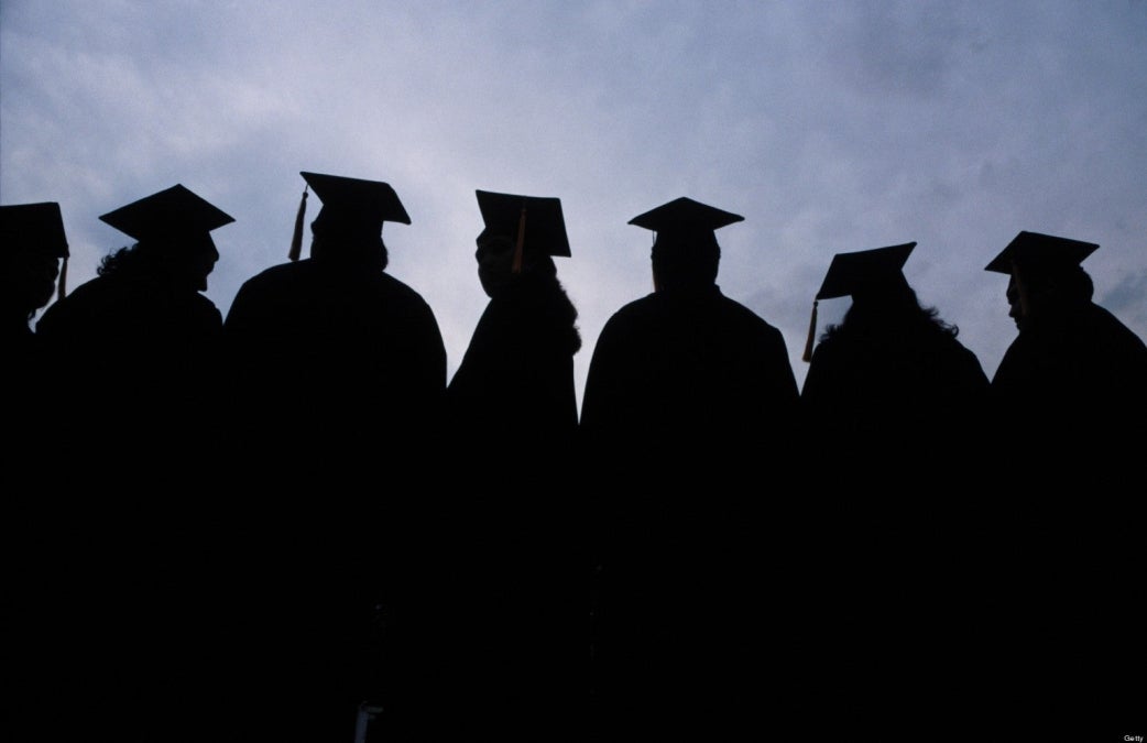 A group of graduates wearing robes, mortar boards and tassles stand in shadow against a blue sky. 