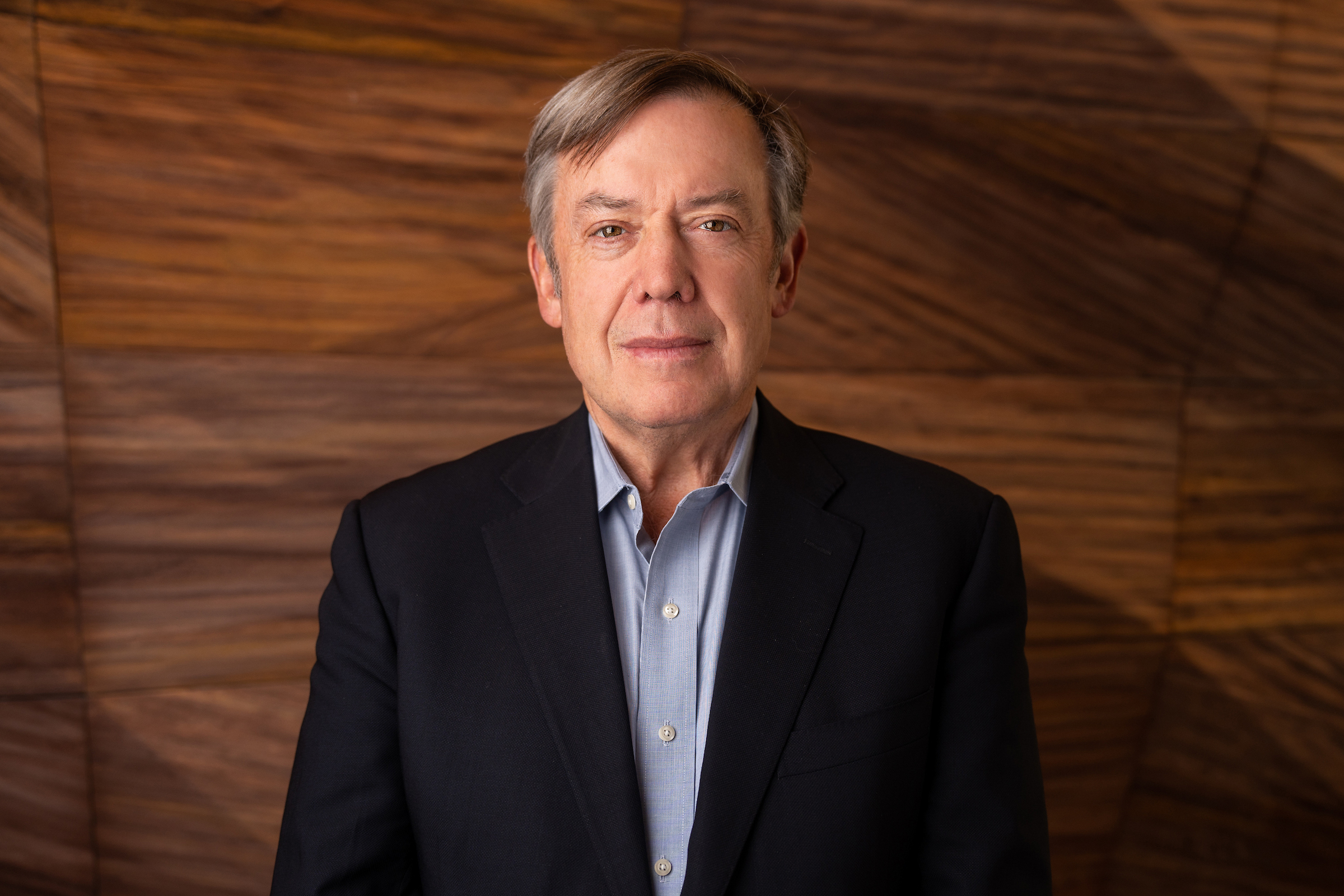 A person in a blue shirt and jacket poses for a headshot in front of a wood paneled wall.