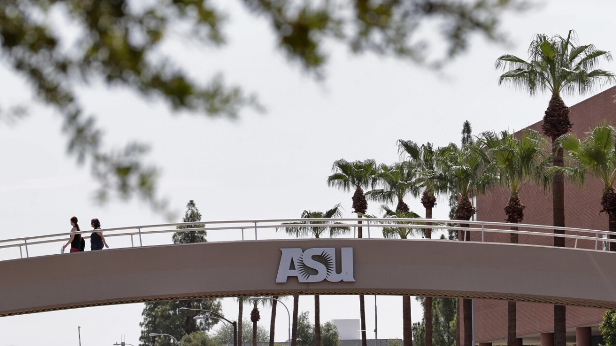 Three people walk across a pedestrian bridge featuring the ASU logo over a street in Tempe, Arizona.