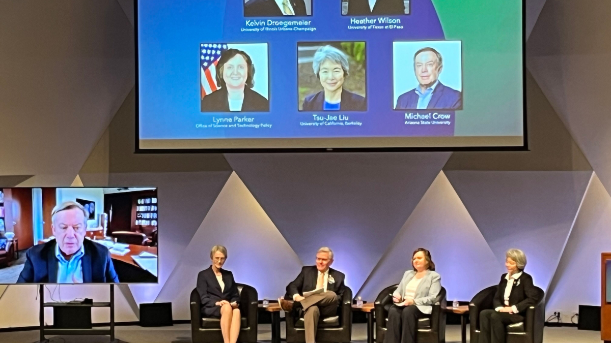 Four science policy and education leaders sit on-stage during the 2025 NAS State of Science address while a fifth speakers shares remarks from a portable screen.