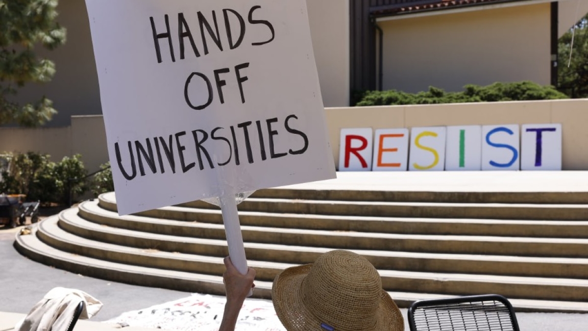 A person holds a protest sign that reads "Hands off universities."